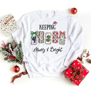 Può includere: Felpa bianca con la scritta "KEEPING Merry & Bright" e grafiche di confezioni regalo festive. Il design include un cappello di Babbo Natale e i numeri 4, 6, 3, 9 e 1. Regali e ornamenti a tema natalizio circondano la felpa.
