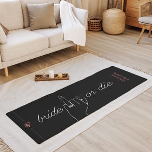 Könnte beinhalten: Schwarze Yogamatte mit den Worten "bride or die" in weißer Schrift. Eine Hand mit einem Ring am Ringfinger ist weiß dargestellt. Die Matte enthält auch den Text "Rachel's Bach June 5, 2023" in Rosa.