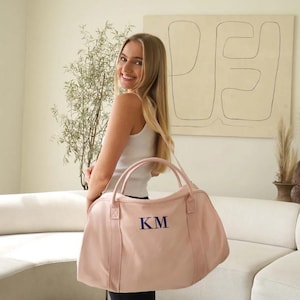 May include: A light pink duffel bag with navy blue embroidered initials "KM". The bag has two handles and a shoulder strap. The bag is made of a soft material and is ideal for travel or the gym.