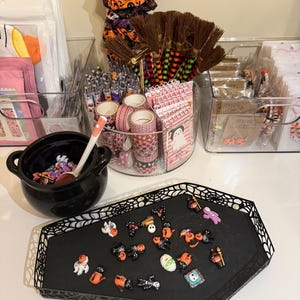 May include: A Halloween-themed display featuring a black coffin-shaped tray with various miniature decorations, a black cauldron with a spoon, and clear containers filled with craft supplies like pens, washi tape, and small brooms. The decorations include ghosts, bats, and pumpkins.
