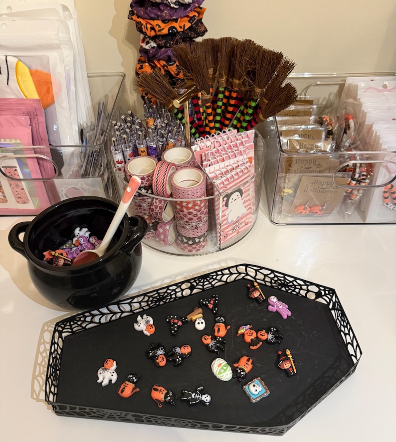May include: A Halloween-themed display featuring a black coffin-shaped tray with various miniature decorations, a black cauldron with a spoon, and clear containers filled with craft supplies like pens, washi tape, and small brooms. The decorations include ghosts, bats, and pumpkins.