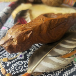 May include: A carved wooden pipe, resembling an animal head, with a carved pattern on top. A feather with brown and white stripes and an orange tip lies near the pipe. The pipe is brown and rests on patterned fabric. The pipe is likely for smoking.