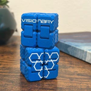 Op de afbeelding: Een blauwe fidget cube met het woord "VISIONARY" in het wit gedrukt. De kubus is gemaakt van verschillende verbonden blokken, met witte geometrische ontwerpen op het onderste gedeelte. De kubus staat op een houten oppervlak.