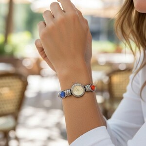 Peut inclure: Une montre analogique argentée dotée d'un cadran rond couleur crème, d'un bracelet métallique et de deux ornements sphériques décoratifs bleu et orange placés de chaque côté du boîtier, fixée par un mécanisme de fermoir simple.