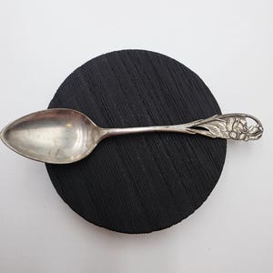 Può includere: Un cucchiaio d'argento con un ornato motivo floreale sul manico è appoggiato su un sottobicchiere nero e rotondo. La coppetta del cucchiaio è ovale e il manico presenta intagli dettagliati di fiori e foglie.