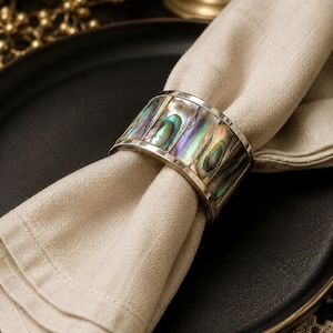 May include: A cylindrical napkin ring featuring a metallic frame inlaid with vertical segments of iridescent abalone shell. The shell displays a spectrum of green, blue, and purple hues with a polished finish, encircling a folded beige linen napkin.