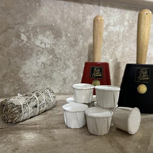 May include: A collection of items including a bundle of white sage, several small white paper cups, and two cowbells. One cowbell is red, the other is black, both with wooden handles and gold accents.
