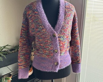 Può includere: Un cardigan multicolore con scollo a V e finiture viola sfocate. Il maglione presenta un motivo a maglia strutturato con tonalità di rosso, arancione, giallo, verde e blu. Anche i polsini e l'orlo sono viola.