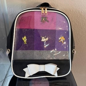 Puede incluir: Mochila negra con un lazo blanco y un panel frontal transparente. El panel muestra un diseño con bloques de color brillante en rosa, morado y gris. Cremalleras doradas y alfileres de personajes de dibujos animados son visibles.
