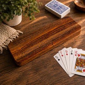 May include: A wooden cribbage board with drilled holes and track lines, alongside a deck of cards and a few fanned-out playing cards. The board is a warm brown color and sits on a wooden surface. A small plant in a white pot is in the background.