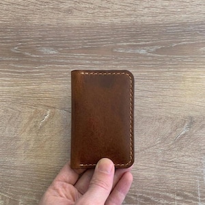 Puede incluir: Un tarjetero rectangular de cuero marr&oacute;n con esquinas redondeadas y costuras visibles de color claro a lo largo de los bordes. El cuero tiene una textura suave y mate con variaciones naturales en el tono marr&oacute;n.