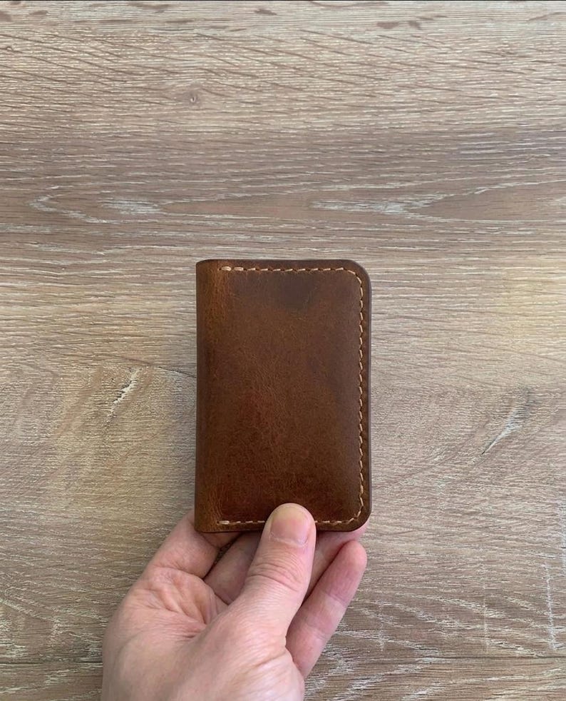 Puede incluir: Un tarjetero rectangular de cuero marr&oacute;n con esquinas redondeadas y costuras visibles de color claro a lo largo de los bordes. El cuero tiene una textura suave y mate con variaciones naturales en el tono marr&oacute;n.