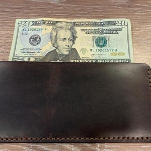 May include: A dark brown leather wallet with a twenty-dollar bill partially inserted. The wallet is rectangular with stitched edges. The bill features a portrait of Andrew Jackson.
