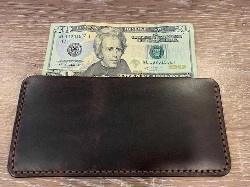 May include: A dark brown leather wallet with a twenty-dollar bill partially inserted. The wallet is rectangular with stitched edges. The bill features a portrait of Andrew Jackson.