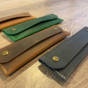 May include: Four rectangular leather cases with rounded flaps, each secured by a single brass-colored metal snap button. The cases are presented in four distinct colors: light tan, forest green, medium brown, and dark charcoal gray.