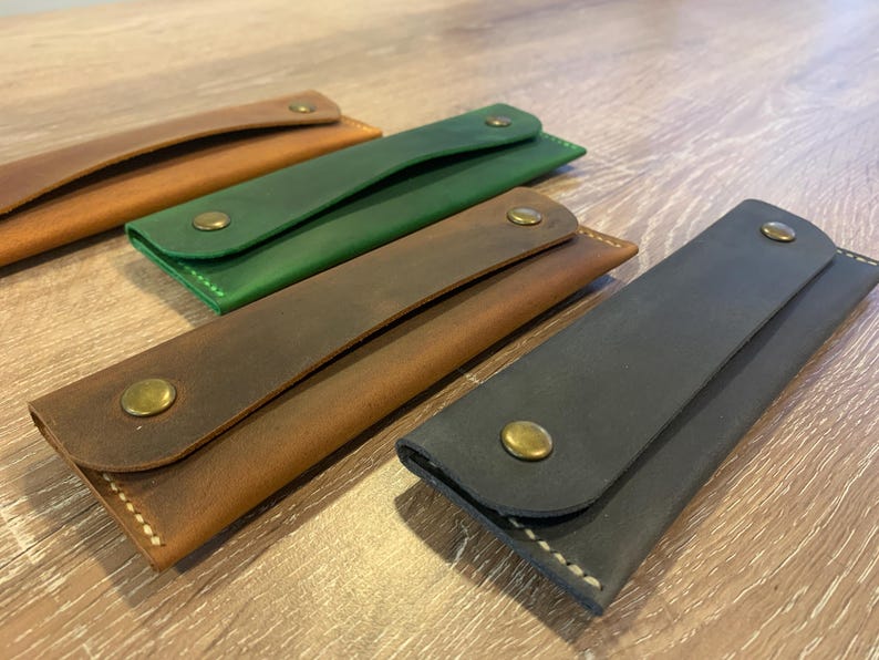 May include: Four rectangular leather cases with rounded flaps, each secured by a single brass-colored metal snap button. The cases are presented in four distinct colors: light tan, forest green, medium brown, and dark charcoal gray.