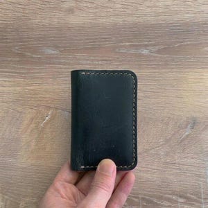 Puede incluir: Un tarjetero rectangular de cuero negro con esquinas redondeadas y costuras visibles de color claro a lo largo de los bordes. La superficie del cuero muestra una textura natural con variaciones sutiles y ligeras marcas de desgaste en el frente.