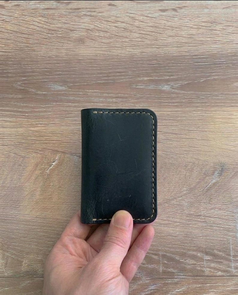 Puede incluir: Un tarjetero rectangular de cuero negro con esquinas redondeadas y costuras visibles de color claro a lo largo de los bordes. La superficie del cuero muestra una textura natural con variaciones sutiles y ligeras marcas de desgaste en el frente.