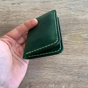 Puede incluir: Un tarjetero de cuero verde oscuro con acabado liso y bordes en contraste cosidos a mano en color verde claro. El accesorio rectangular tiene un dise&ntilde;o plegado con costuras visibles a lo largo del per&iacute;metro en un tono esmeralda intenso.