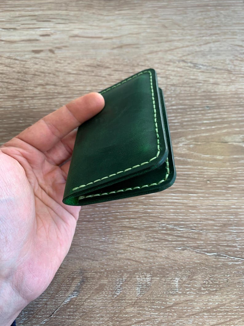 Puede incluir: Un tarjetero de cuero verde oscuro con acabado liso y bordes en contraste cosidos a mano en color verde claro. El accesorio rectangular tiene un dise&ntilde;o plegado con costuras visibles a lo largo del per&iacute;metro en un tono esmeralda intenso.
