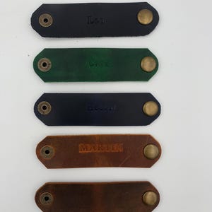 Peut inclure: Cinq lanières en cuir rectangulaires aux coins arrondis, chacune dotée d'un bouton-pression en métal couleur laiton aux deux extrémités. Les lanières sont noires, vert foncé, bleu marine et deux tons de marron. Trois lanières portent les noms Leo, Carol, Hector et MARTIN.