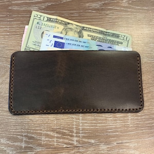 May include: A dark brown leather wallet with a rectangular shape. The wallet is filled with a twenty-dollar bill and a blue and white bill. The wallet has a stitched border and is placed on a wooden surface.