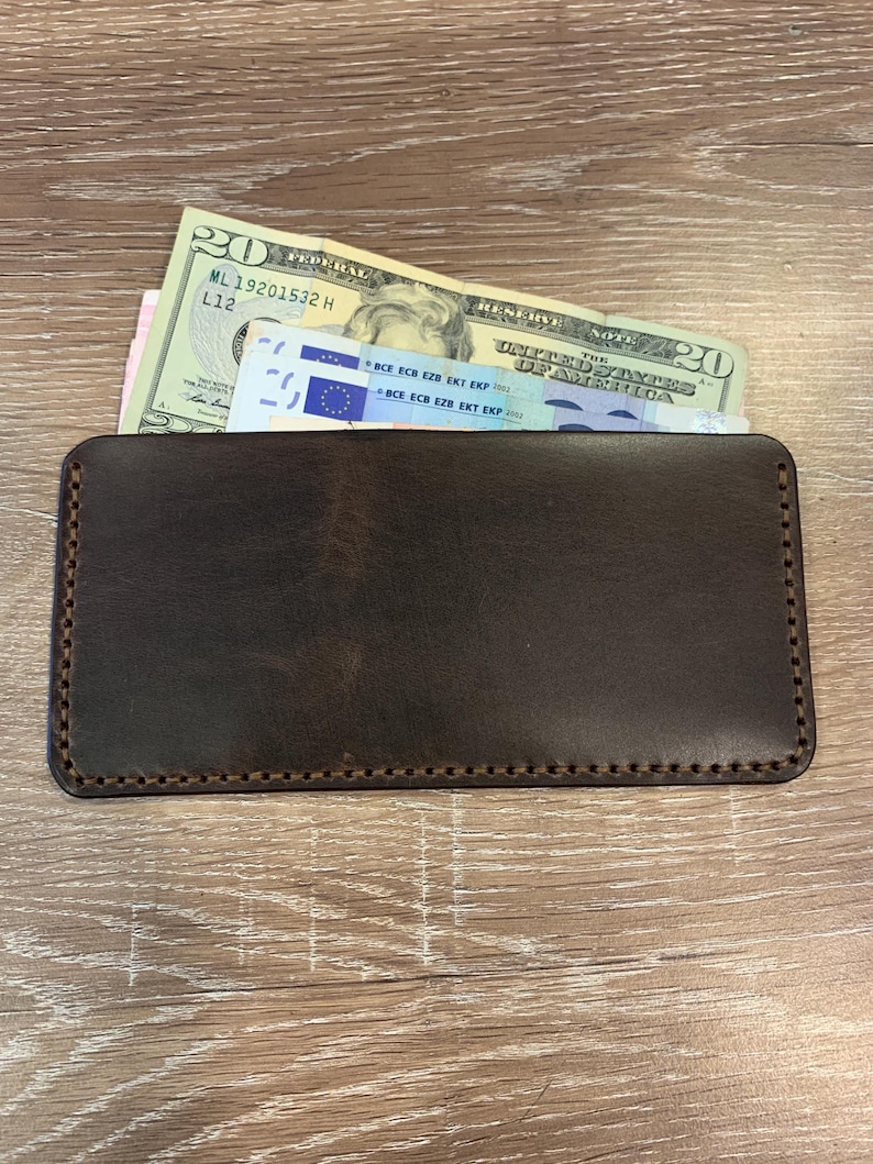 May include: A dark brown leather wallet with a rectangular shape. The wallet is filled with a twenty-dollar bill and a blue and white bill. The wallet has a stitched border and is placed on a wooden surface.