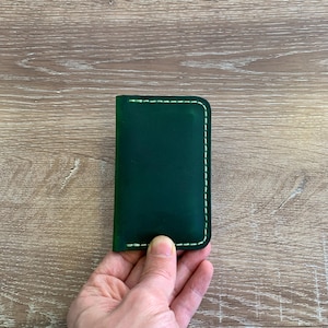 Puede incluir: Un tarjetero rectangular de cuero verde oscuro con esquinas redondeadas y costuras de color claro a lo largo de los bordes. El art&iacute;culo tiene un acabado liso y mate, con orientaci&oacute;n vertical para guardar tarjetas o billetes doblados.