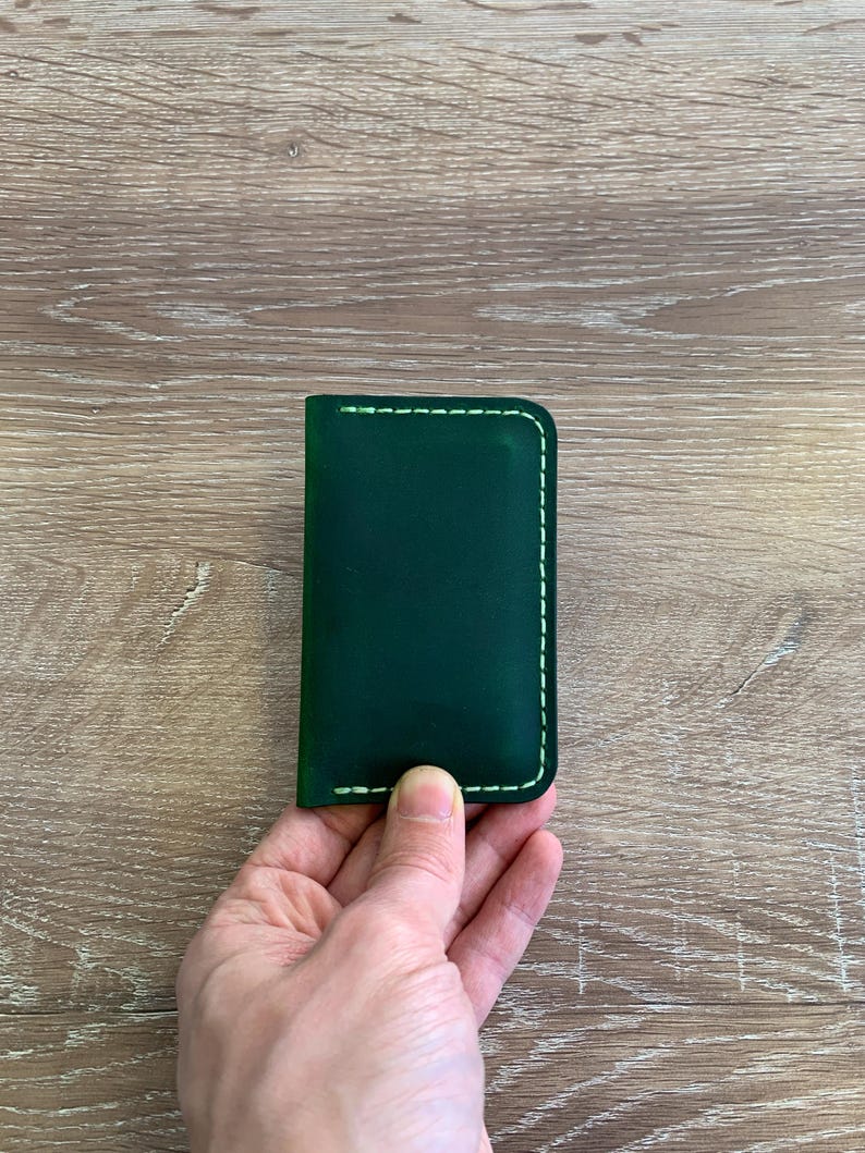 Puede incluir: Un tarjetero rectangular de cuero verde oscuro con esquinas redondeadas y costuras de color claro a lo largo de los bordes. El art&iacute;culo tiene un acabado liso y mate, con orientaci&oacute;n vertical para guardar tarjetas o billetes doblados.