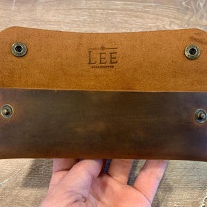 May include: A rectangular brown leather wallet featuring a fold-over flap with two metal snap buttons. The interior displays the embossed text LEE HANDCRAFTED with a small star icon. The exterior is stitched with light-colored thread at the sides.