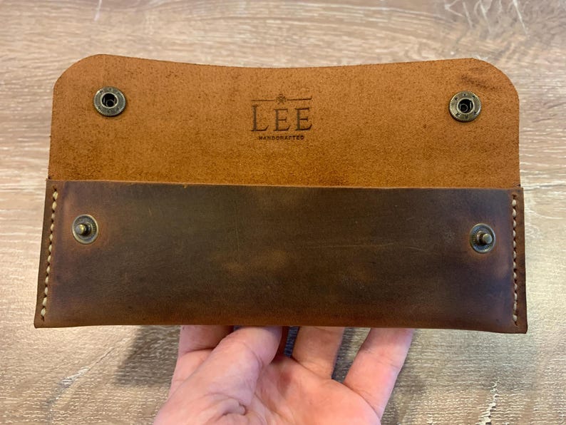 May include: A rectangular brown leather wallet featuring a fold-over flap with two metal snap buttons. The interior displays the embossed text LEE HANDCRAFTED with a small star icon. The exterior is stitched with light-colored thread at the sides.