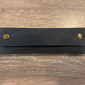 May include: A rectangular black leather case featuring a fold-over flap secured by two brass-colored metal snap buttons. The edges of the case are joined with visible light-colored stitching, creating a clean and functional design for storage.