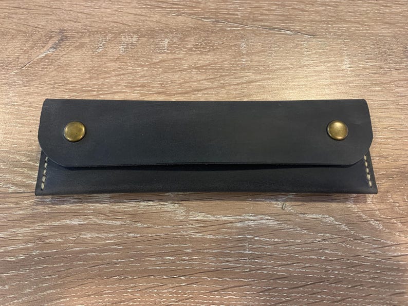 May include: A rectangular black leather case featuring a fold-over flap secured by two brass-colored metal snap buttons. The edges of the case are joined with visible light-colored stitching, creating a clean and functional design for storage.