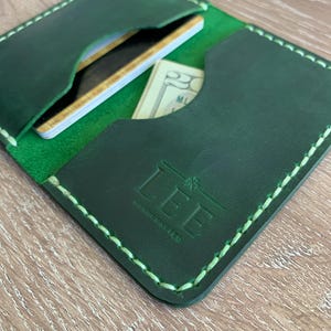 Puede incluir: Un tarjetero de cuero verde oscuro con costuras de color verde claro a lo largo de los bordes. La parte frontal tiene grabado el texto LEE HANDCRAFTED. El interior contiene varias ranuras con tarjetas de cr&eacute;dito y un billete de veinte d&oacute;lares.
