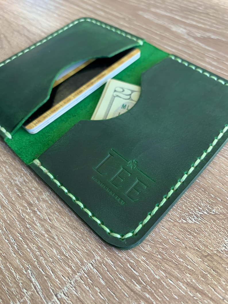 Puede incluir: Un tarjetero de cuero verde oscuro con costuras de color verde claro a lo largo de los bordes. La parte frontal tiene grabado el texto LEE HANDCRAFTED. El interior contiene varias ranuras con tarjetas de cr&eacute;dito y un billete de veinte d&oacute;lares.