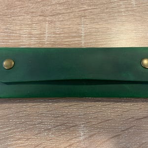 May include: A rectangular forest green leather case featuring a fold-over flap secured by two brass-colored metal snap buttons. The edges are stitched with matching green thread, creating a clean and functional design for storing small items.