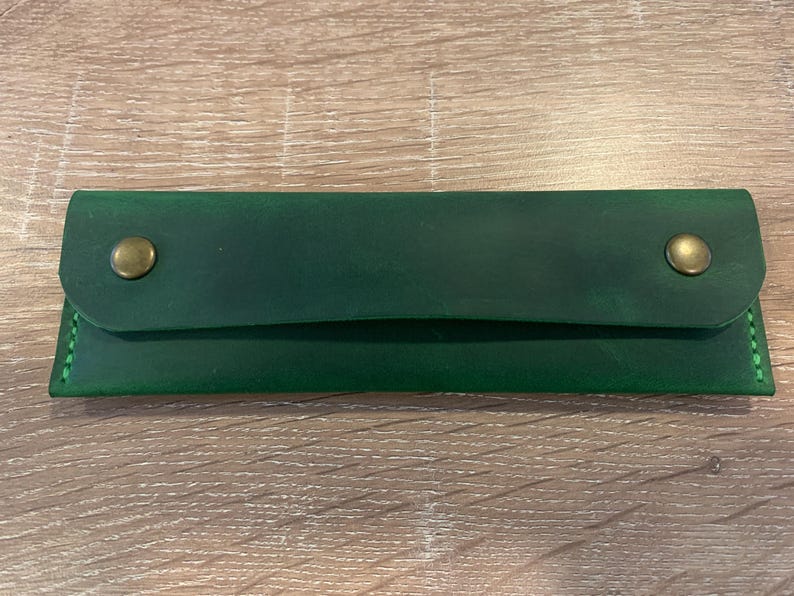 May include: A rectangular forest green leather case featuring a fold-over flap secured by two brass-colored metal snap buttons. The edges are stitched with matching green thread, creating a clean and functional design for storing small items.