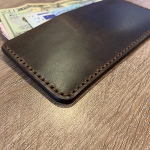 May include: A dark brown leather wallet with visible stitching around the edges. The wallet is rectangular and appears to be filled with cash and possibly other documents. The leather has a smooth, polished finish.