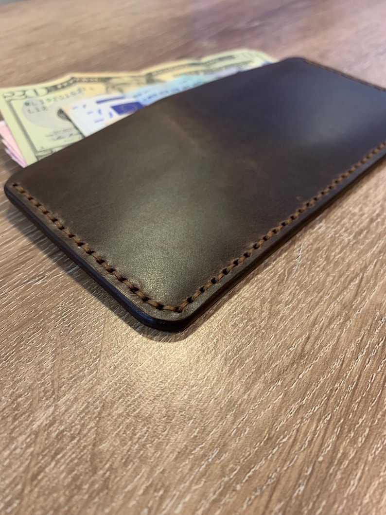 May include: A dark brown leather wallet with visible stitching around the edges. The wallet is rectangular and appears to be filled with cash and possibly other documents. The leather has a smooth, polished finish.