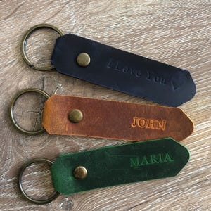 May include: Three leather keychains with brass-toned metal rings and rivets. The top keychain is black with the text "I Love You" and a heart symbol. The middle keychain is brown with the name "JOHN" embossed. The bottom keychain is green with the name "MARIA" embossed.