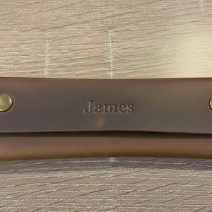 May include: A rectangular brown leather case featuring a flap closure secured by two brass-colored metal snaps. The name James is embossed in the center of the flap. The case has visible light-colored stitching along the side edges.