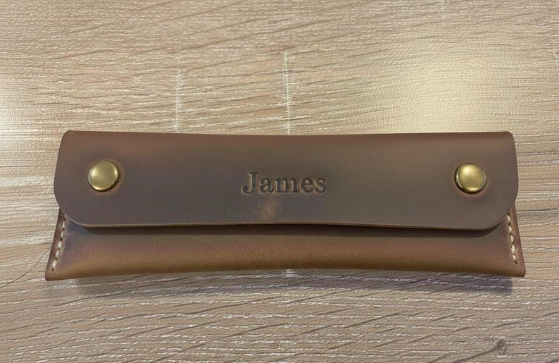 May include: A rectangular brown leather case featuring a flap closure secured by two brass-colored metal snaps. The name James is embossed in the center of the flap. The case has visible light-colored stitching along the side edges.