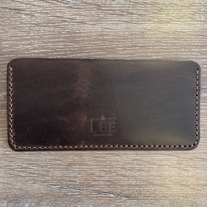 May include: A dark brown leather wallet with a rectangular shape. The wallet has a stitched border and the word "LEE" is embossed on the front. The wallet is shown on a light brown wooden surface.