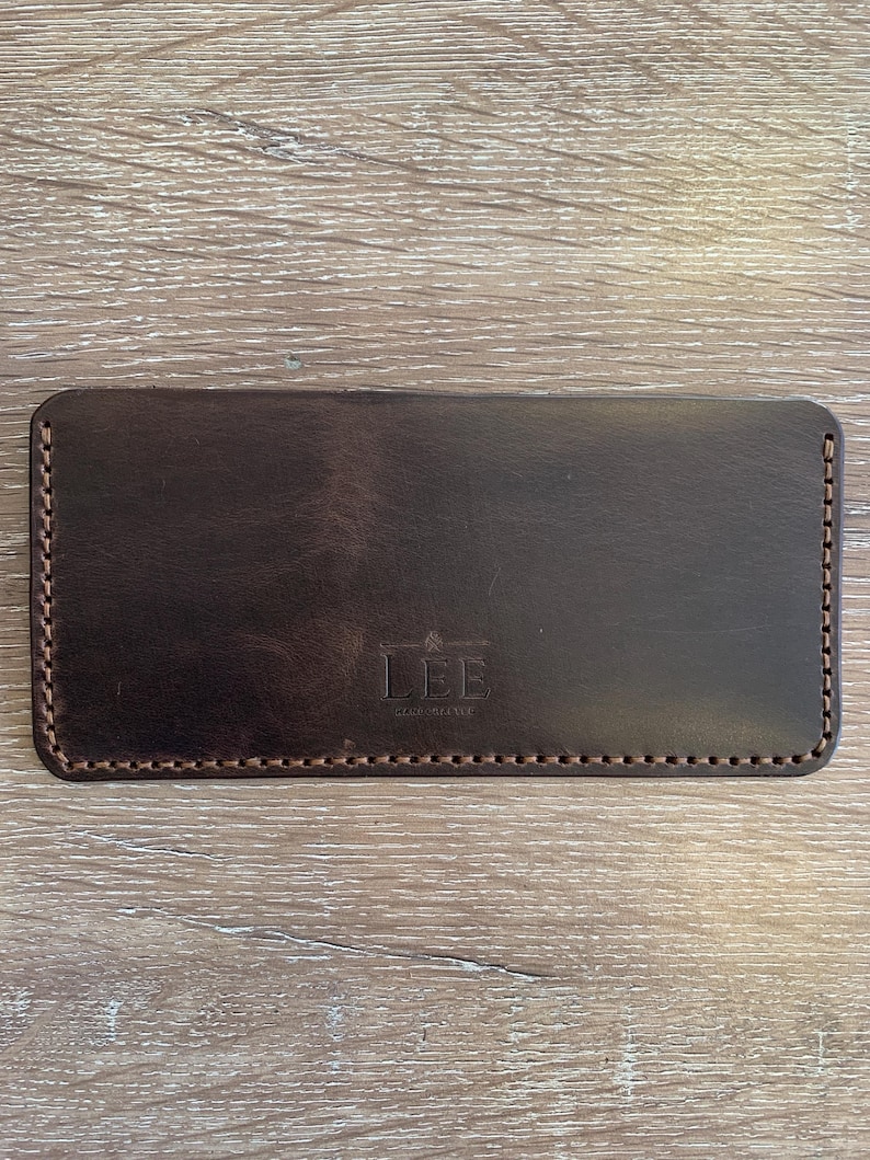 May include: A dark brown leather wallet with a rectangular shape. The wallet has a stitched border and the word "LEE" is embossed on the front. The wallet is shown on a light brown wooden surface.