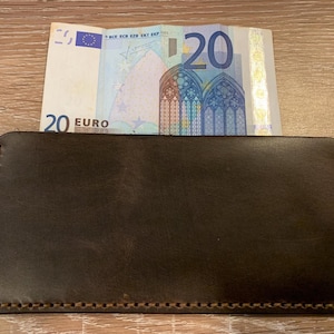 May include: A dark brown leather wallet with a 20 Euro bill partially inserted. The wallet is rectangular with stitched edges. The bill is visible, displaying the number 20 and architectural details.