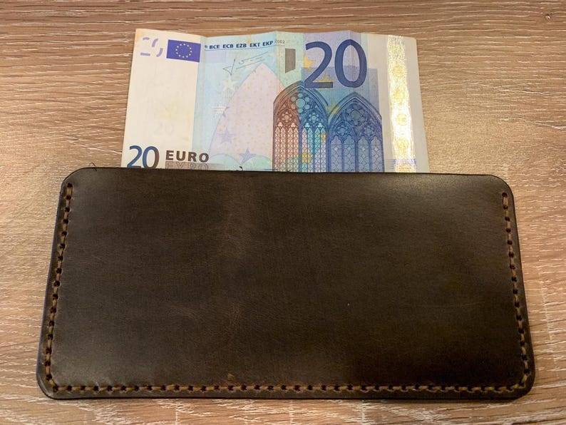 May include: A dark brown leather wallet with a 20 Euro bill partially inserted. The wallet is rectangular with stitched edges. The bill is visible, displaying the number 20 and architectural details.