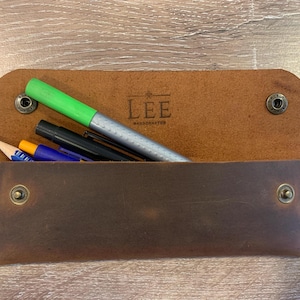 May include: A brown leather pencil case with a flap closure secured by two metal snaps. The interior is stamped with the text LEE HANDCRAFTED. Inside the case are several writing instruments, including a bright green marker and a blue pen.