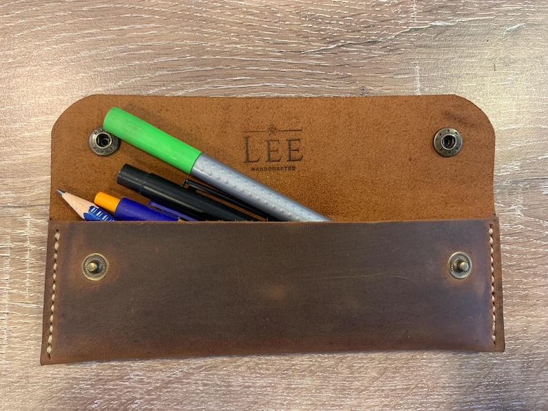 May include: A brown leather pencil case with a flap closure secured by two metal snaps. The interior is stamped with the text LEE HANDCRAFTED. Inside the case are several writing instruments, including a bright green marker and a blue pen.
