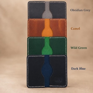 Puede incluir: Una pila de cuatro tarjeteros rectangulares en gris obsidiana, camel, verde salvaje y azul oscuro. Cada tarjetero presenta una muesca central para el pulgar y costuras blancas en contraste. El tarjetero azul oscuro tiene el texto LEE.