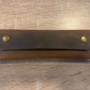 May include: A rectangular brown leather case with a fold-over flap secured by two brass-colored metal snap buttons. The leather features visible stitching along the side edges and a smooth, matte finish with natural variations in the brown tone.